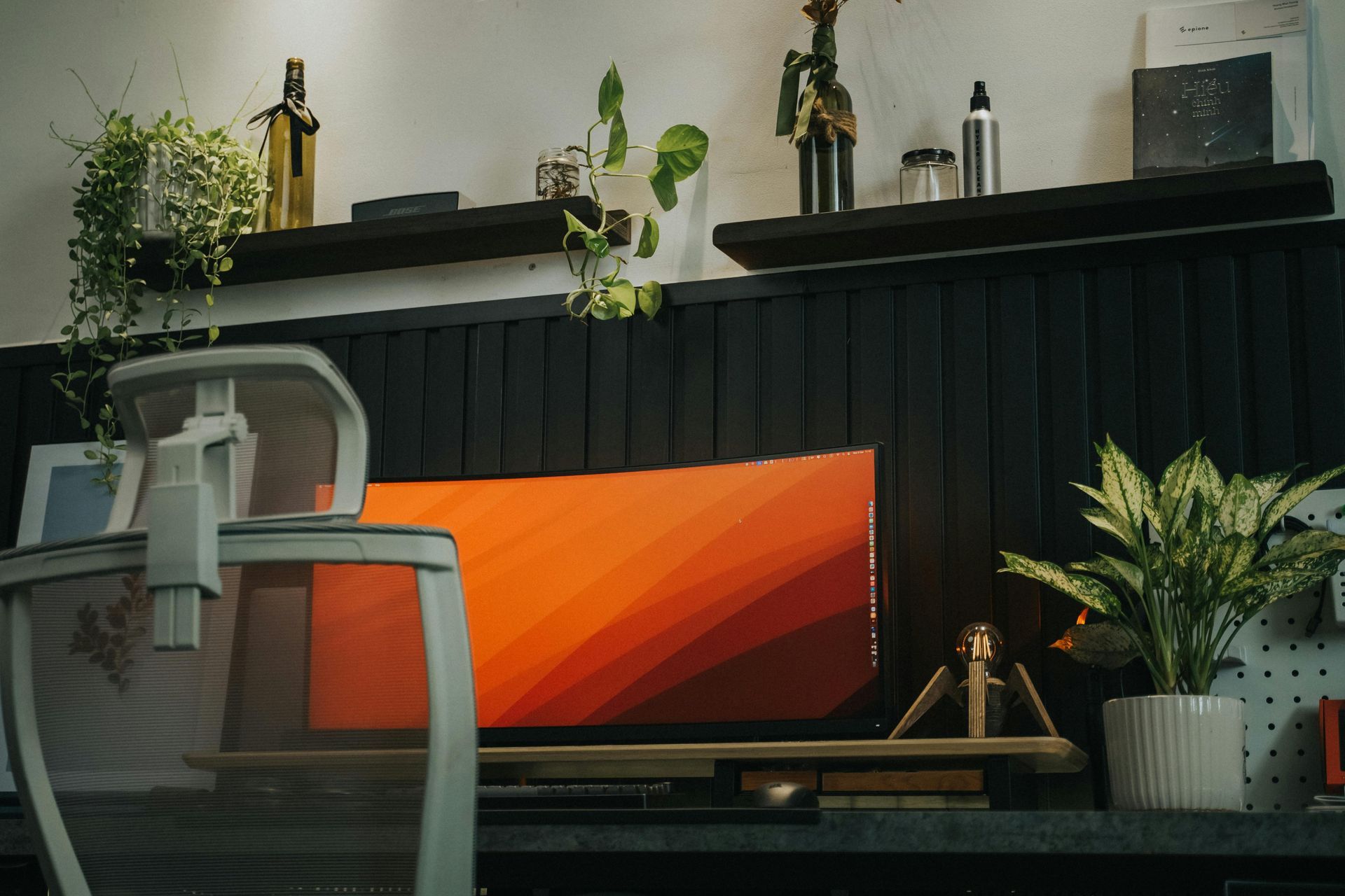 Modern desk setup with orange monitor, white chair, black shelf, and potted plants on a dark desk